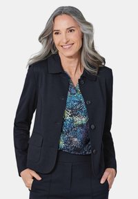 Zwarte op maat gemaakte blazer met een kraag, voorzien van vier knopen en twee voorzakken. Gedragen over een blouse met een patroon in blauw en groen.