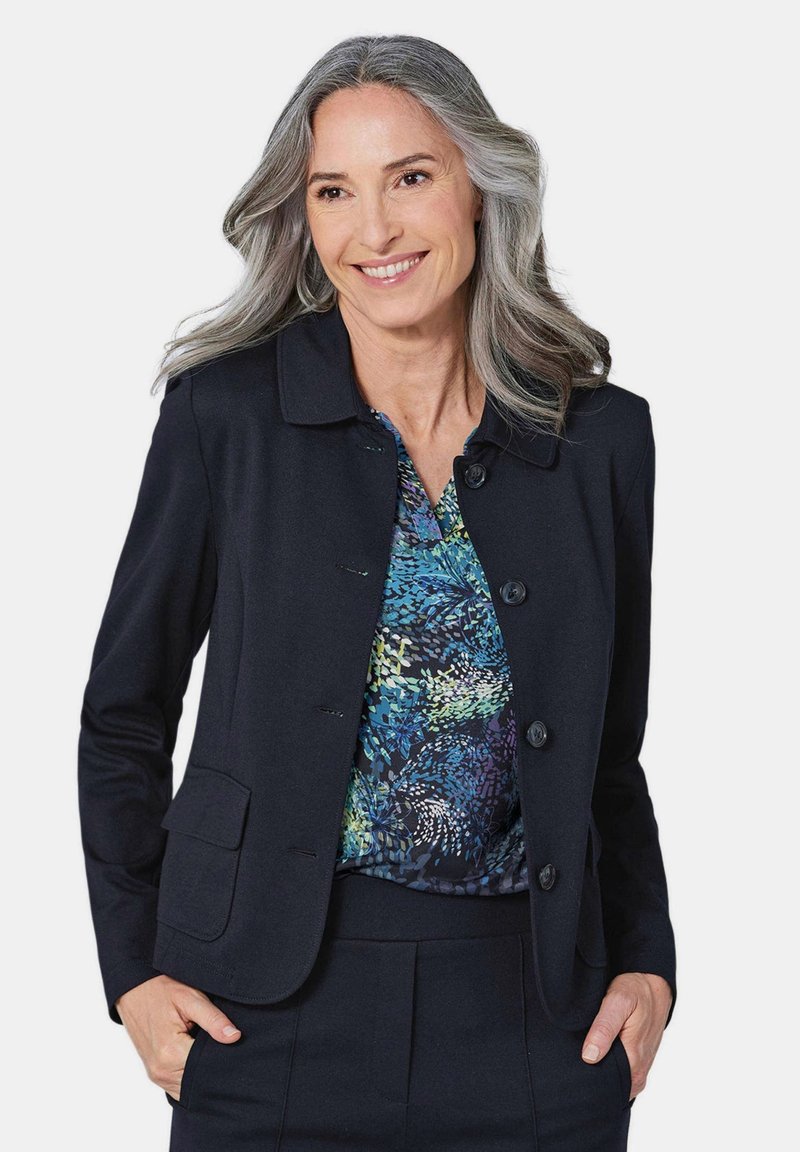 Zwarte op maat gemaakte blazer met een kraag, voorzien van vier knopen en twee voorzakken. Gedragen over een blouse met een patroon in blauw en groen.