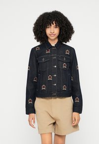 Tmavomodrá rifľová bunda s vyšívanými motívmi koruny v červenej a zlatej farbe, s gombíkovým zapínaním vpredu a dvoma náprsnými vreckami. Nosená s béžovými šortkami.