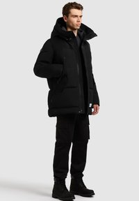 Juoda pūkinė striukė su gobtuvu, pasiūta su kviltuoto dizaino, priekyje užtrauktuku ir šoniniais kišenėmis, derinama su juodomis karo stiliaus kelnėmis ir batais.