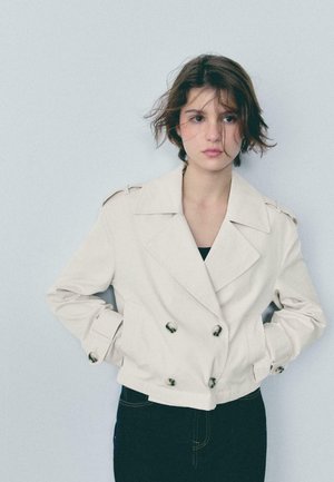 Jonge persoon met kort, warrig haar draagt een lichtbeige dubbelgeknoopte jas en donkere broek, staand tegen een effen lichtgrijze achtergrond.