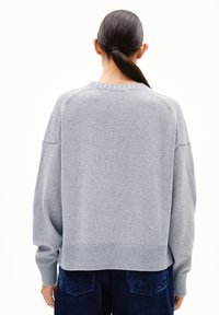 Frau mit schwarzen Haaren in einem tiefen Pferdeschwanz, die einen lockeren hellgrauen Pullover und dunkle blaue Cordhosen trägt, und von einem weißen Hintergrund abgewandt ist.