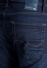 Temno modri kavbojski hlače z značilnim pet-žepnim dizajnom, z belim šivom in usnjenim oznako na zadnjem pasu.