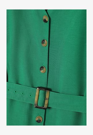 Zelené šaty s textúrovanou tkaninou, s veľkými okrúhlymi gombíkmi a zodpovedajúcim opaskom so štvorcovou prackou. Ozdobené prešívaním.