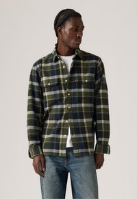 Groene en marineblauwe geruite flanellen shirt met twee borstzakken en knoopsluitingen, gecombineerd met een wit t-shirt en blauwe spijkerbroek.