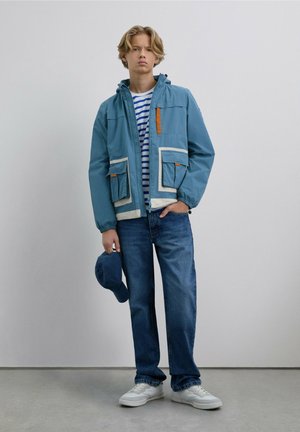 Chaqueta de color azul claro con ribetes blancos y acento naranja, con dos bolsillos frontales, llevada sobre una camiseta a rayas. Combinada con jeans azules y zapatillas blancas.