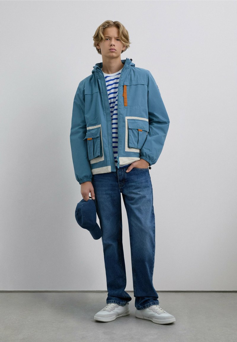 Chaqueta de color azul claro con ribetes blancos y acento naranja, con dos bolsillos frontales, llevada sobre una camiseta a rayas. Combinada con jeans azules y zapatillas blancas.