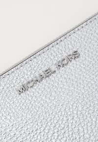 Novčanik od srebrne kožne strukture s teksturiranom površinom, metalnim dijelovima u srebrnoj boji i utisnutim "Michael Kors" logotipom na prednjoj strani.