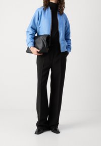 Chaqueta bomber de color azul claro con un patrón texturizado, cuello de tortuga negro, pantalones negros a medida y un bolso negro informal. Zapatos negros.