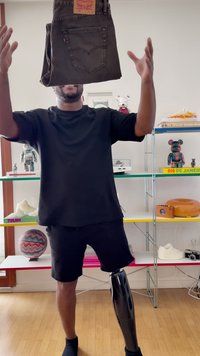 Moški s protetično nogo meče temne kavbojke pred pisano policjo s knjigami, igračami in športnimi čevlji.