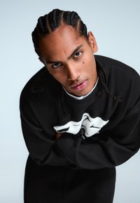 Čierny pletený sveter s bielym grafickým dizajnom, s okrúhlym výstrihom a dlhými rukávmi. Model má zapletané vlasy a prekrížené ruky.