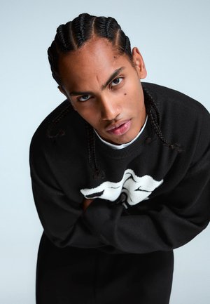 Schwarzer Strickpullover mit weißem Grafikdesign, Rundhals und langen Ärmeln. Das Model hat geflochtenes Haar und verschränkte Arme.