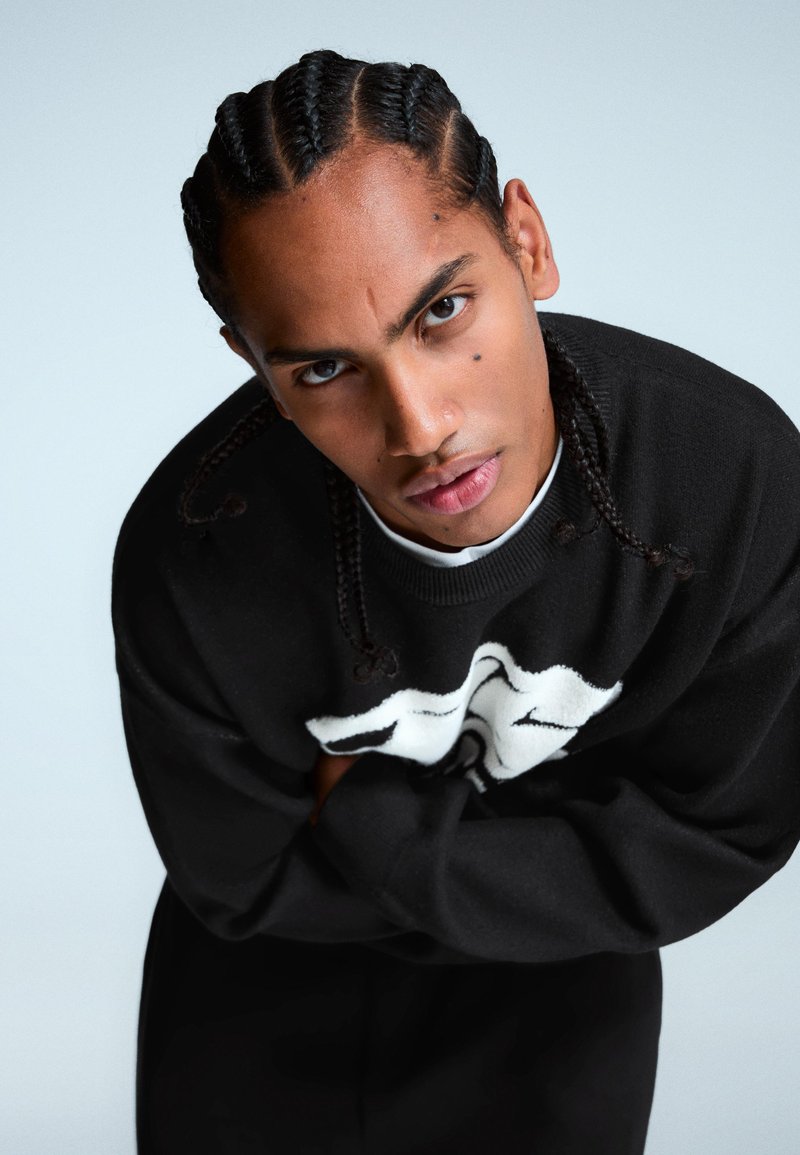 Čierny pletený sveter s bielym grafickým dizajnom, s okrúhlym výstrihom a dlhými rukávmi. Model má zapletané vlasy a prekrížené ruky.