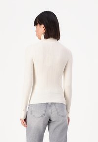 Pull en tricot de couleur crème avec une texture côtelée, un col haut et des accents de boutons sur les poignets. Porté avec un jean gris.