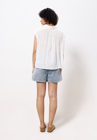 Leichte weiße Bluse mit kurzen Ärmeln und gefälteltem Rückendetail; kombiniert mit Jeansshorts und Schuhen aus hellbraunem Wildleder.