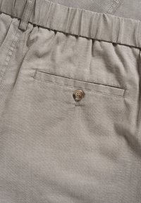 Los pantalones de lino beige cuentan con una cinturilla elástica y un único bolsillo delantero con un cierre de botón marrón, resaltando una textura suave.