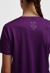 Lila sporttröja med mjuk textur och korta ärmar, med en vit chevrondesign på ryggen nära halsringningen.