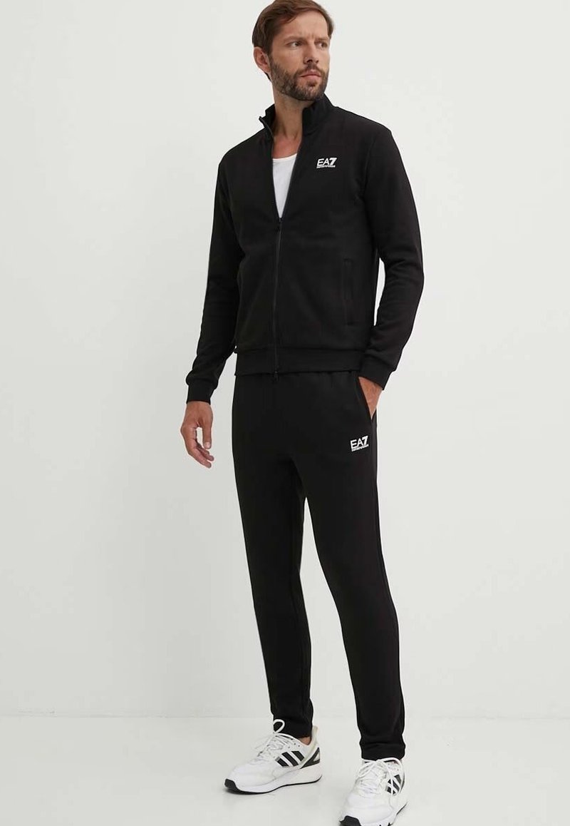 Tuta nera realizzata in morbido tessuto; giacca con zip e alta collana, pantaloni con vita elastica; presenta il logo "EAZ" in bianco.