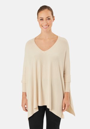 Smilende kvinde iført løs, beige V-hals trøje med asymmetrisk kant og sorte bukser, stående mod en hvid baggrund.