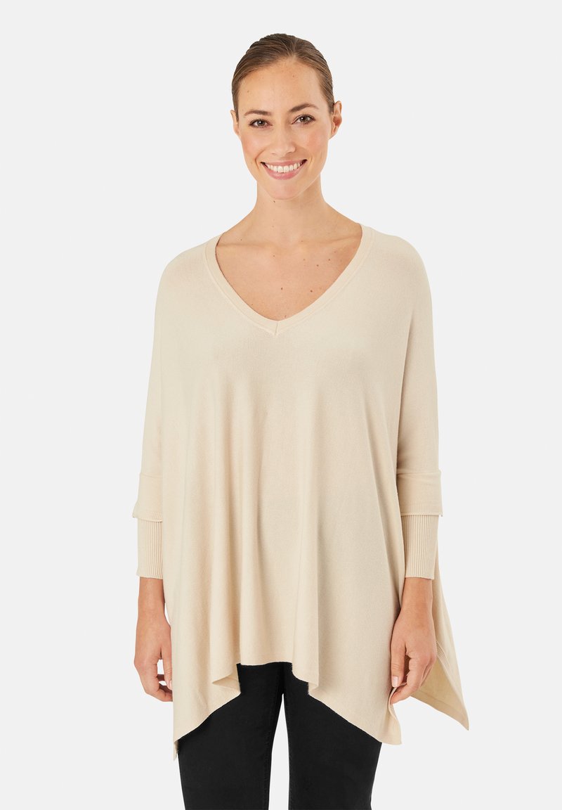 Lächelnde Frau, die einen lockeren, beigefarbenen V-Ausschnittpullover mit asymmetrischem Saum und schwarze Hose trägt, vor einem weißen Hintergrund steht.