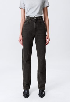 Personne portant un jean noir taille haute associé à une chemise gris clair à manches courtes rentrée et des chaussures noires à bout pointu.