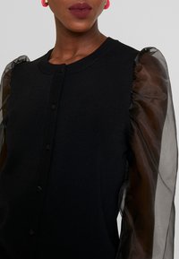 Cárdigan negro con cierre de botones, con mangas abullonadas transparentes en negro. Textura suave y cuerpo ajustado.