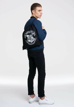 Black drawstring bag featuring a white Hogwarts crest design, made of fabric, worn by a person with a navy bomber jacket and black pants.