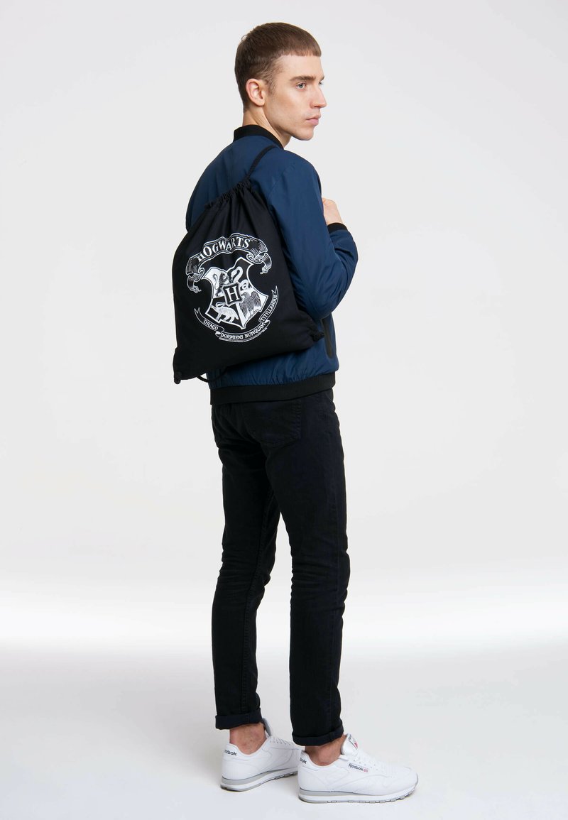 Black drawstring bag featuring a white Hogwarts crest design, made of fabric, worn by a person with a navy bomber jacket and black pants.