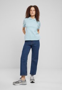 Lyseblå polo med klassisk krage, kombinert med høy midje mørkeblå jeans og grå treningssko. Ensfarget bakgrunn.