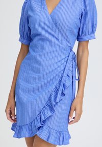Robe portefeuille bleue à rayures verticales blanches, manches ballon, bordure à volants et fermeture à nouer portée par une personne debout aux bras découverts.