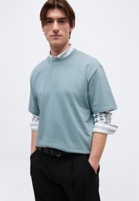 Νέος άνδρας με καστανά μαλλιά που φοράει ανοιχτό μπλε κοντομάνικο πουκάμισο πάνω από ριγωτό μακρυμάνικο πουκάμισο και μαύρο παντελόνι με λεπτές ρίγες.