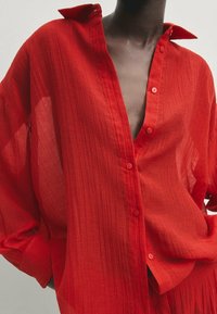 Chemise rouge texturée à boutons avec une coupe ample, présentant un col en V profond, des manches longues et un col plié.