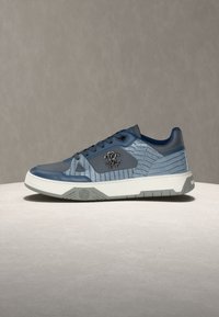 Μπλε και γκρι αθλητικό παπούτσι με υφασμάτινα πάνελ, μεταλλικό λογότυπο στο πλάι, λευκή σόλα και γκρι πέλμα, τοποθετημένο σε φωτεινή επιφάνεια.