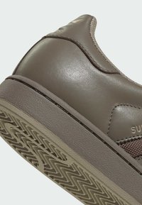 Zapatilla marrón de cuero con superficie texturizada, suela de goma con patrón en zigzag y un detalle de malla en el lateral. Se observa una marca sutil.