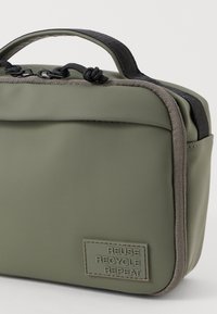 Borsa rettangolare verde con zip nera, manico in tessuto e patch con la scritta "RIUTILIZZA RICICLA RIPETI" nell'angolo in basso a sinistra.