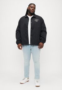 Crna lagana jakna s gumbima sprijeda i elastičnim manžetama, nosi se preko bijele majice i svijetloplavih traperica, s detaljem logotipa na prsima.