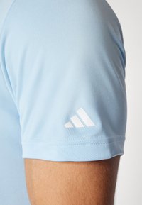 Ljusblå träningsskjorta med korta ärmar, med en vit Adidas-logotyp på övre högra ärmen, slät tygtextur.