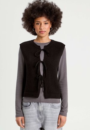 Zwarte gebreide vest met een ronde halslijn, voorzien van drie strikken in het midden. Gecombineerd met een grijs longsleeve-top en lichte jeans.