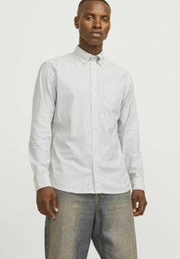 Jasnoszara koszula z długim rękawem, zapinana na guziki, w pionowe białe paski, z kieszenią na piersi, wykonana z gładkiego materiału. Połączona z dżinsami.