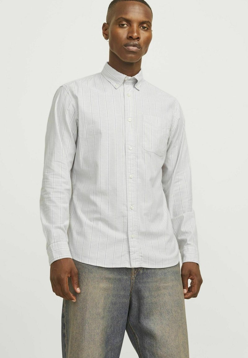 Jasnoszara koszula z długim rękawem, zapinana na guziki, w pionowe białe paski, z kieszenią na piersi, wykonana z gładkiego materiału. Połączona z dżinsami.