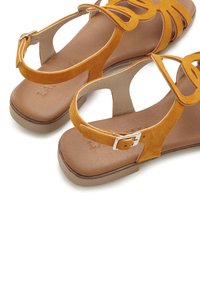 Gelbe Wildleder-Sandalen mit Ausschnittdesign und Knöchelriemen. Flache Sohle mit hellbraunen Akzenten. Schlichte, offene Silhouette.