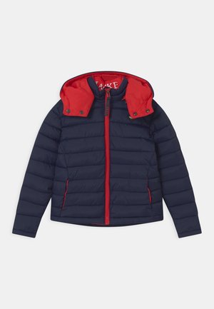 Veste matelassée marine avec une capuche rouge et une fermeture éclair, présentant une texture matelassée horizontale et des accents rouges contrastants le long de la fermeture éclair et des poches.