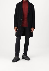 Manteau noir cintré, pull rouge à motifs, jean foncé et chaussures de sport noires avec des accents texturés.