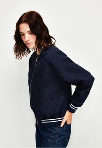 Granatowa kurtka bombowiec z teksturowanej tkaniny, z przednim zamkiem błyskawicznym, białymi paskowanymi mankietami i subtelnym tłoczonym wzorem.