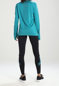 Türkisen Langarm-Sportoberteil mit strukturierten Einsätzen; kombiniert mit schwarzen Leggings, die mit blauen Akzenten und einem Knöchellänge-Design versehen sind.