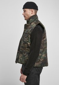 Zöld, barna és fekete camouflázs mintázatú ujjatlan mellény. Magas gallérral és több elülső zsebbel rendelkezik. Sötét kötött pulcsi fölött viselik.