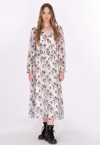Floral jurk tot aan de enkels in een lichte stof, met een diepe V-halslijn, lange doorschijnende mouwen en een smocktail. Gepaarde met zwarte veterschoenen.