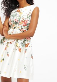 Blommig klänning med vit botten, prydd med färgglada blommönster i olika nyanser. Ärmlös design, A-linjeformad och med knytbart midjeband.