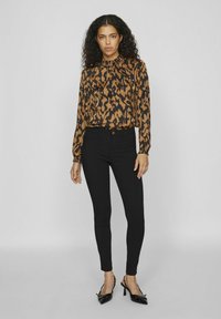 Blouse à manches longues et col, avec un motif abstrait marron et noir, assortie à un pantalon noir ajusté. La tenue présente une texture lisse.