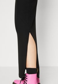 Calças pretas com textura canelada, apresentando uma abertura lateral, combinadas com botas rosa-choque com atacadores e meias pretas. O design enfatiza tanto a cor como a textura.